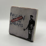 Banksy artwork featuring the phrase 'Follow Your Dreams' printed on square travertine stone coaster