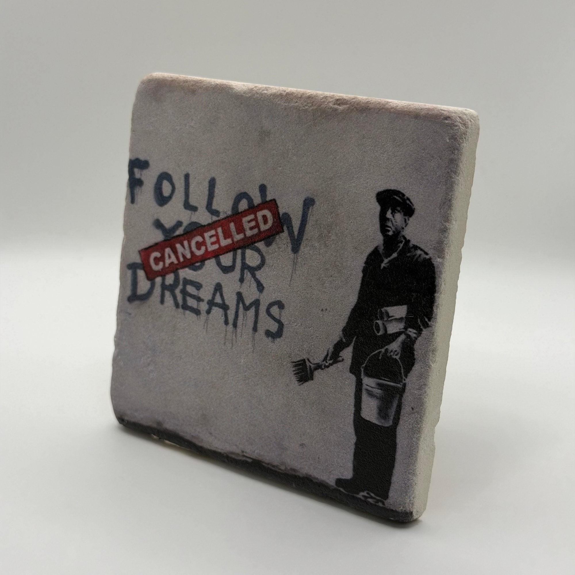 Banksy artwork featuring the phrase 'Follow Your Dreams' printed on square travertine stone coaster