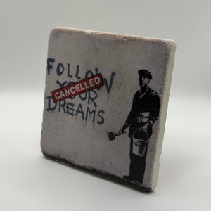 Banksy artwork featuring the phrase 'Follow Your Dreams' printed on square travertine stone coaster